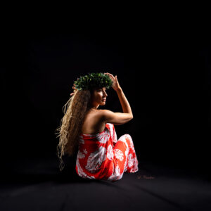 Photo en studio sur fond noir d'une danseuse Tahitienne. Elle est assise avec une couronne de fleur verte et une robe rouge